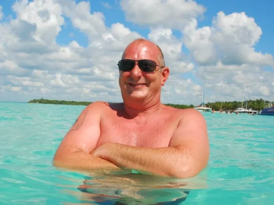 Rainer, lächelnd im türkisfarbenen Wasser, mit Sonnenbrille und einer tropischen Landschaft im Hintergrund.