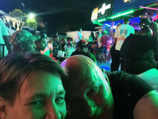 Manuela und Rainer nehmen ein Selfie vor einem belebten Veranstaltungsort (Karaoke) mit bunten Lichtern auf.