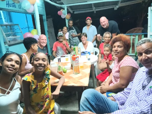 Gruppe von Menschen verschiedenen Alters, darunter Kinder, Erwachsene und ältere Personen, sitzen und stehen um einen Tisch herum bei einer festlichen Veranstaltung im Freien mit Ballons und Getränken